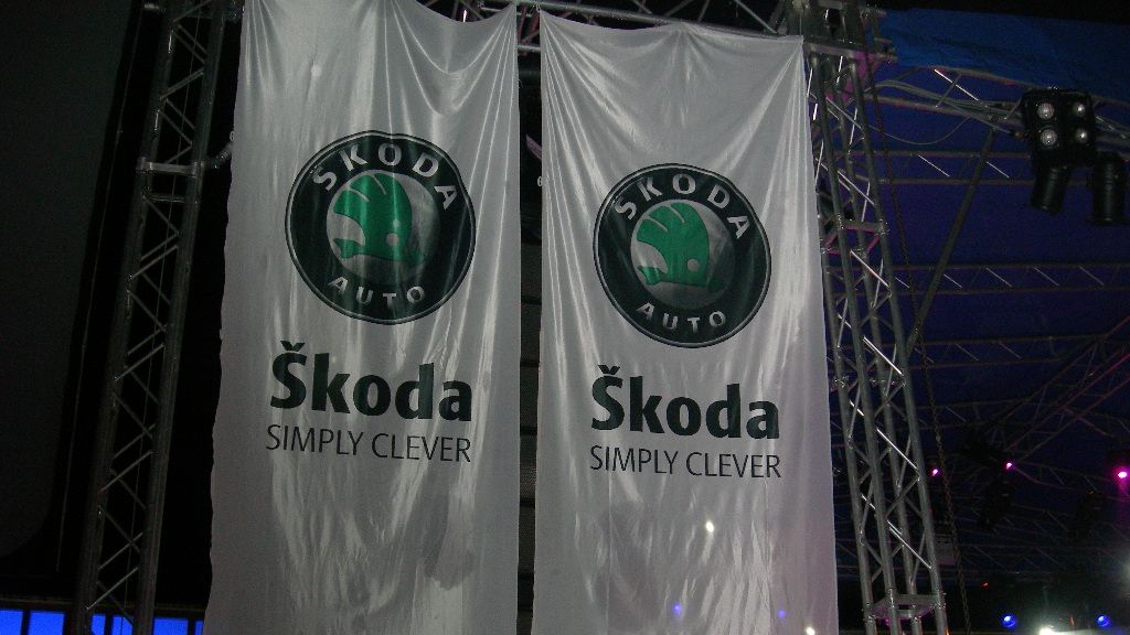 Dy pankarta të mëdha vertikale të bardha varen në një strukturë metalike, secili me logon e gjelbër të Škoda Auto në krye. Nën logo shkruhet “Škoda” me shkronja të mëdha dhe slogani “SIMPLY CLEVER”. Sfondi është një ambient i errët eventesh me ndriçim shumëngjyrësh.